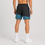 Male athlete wearing black and teal color-block workout shorts, back view, elastic waistband, white socks and sneakers, studio grey background