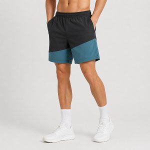 Male model wearing black and teal color-block workout shorts, front view, hands in pockets, white socks and sneakers, studio grey background