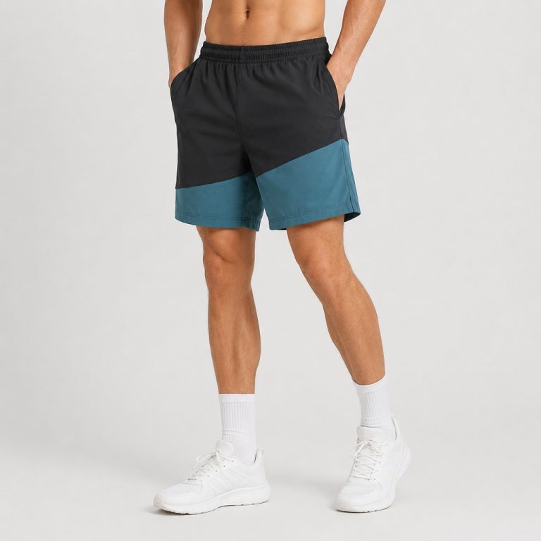 Male model wearing black and teal color-block workout shorts, front view, hands in pockets, white socks and sneakers, studio grey background