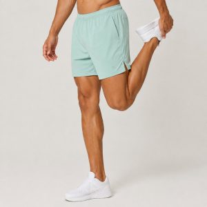 Male athlete wearing mint green workout shorts standing on one leg, stretching quadriceps, white sneakers