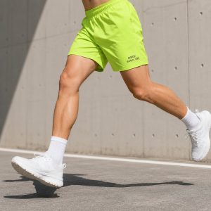 Male athlete running in neon yellow fluorescent athletic shorts with enjoy every day text logo, white socks and sneakers, outdoor concrete wall and asphalt road