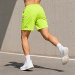 Back and side view of male athlete running in neon yellow fluorescent athletic shorts, white socks and sneakers, outdoor concrete wall