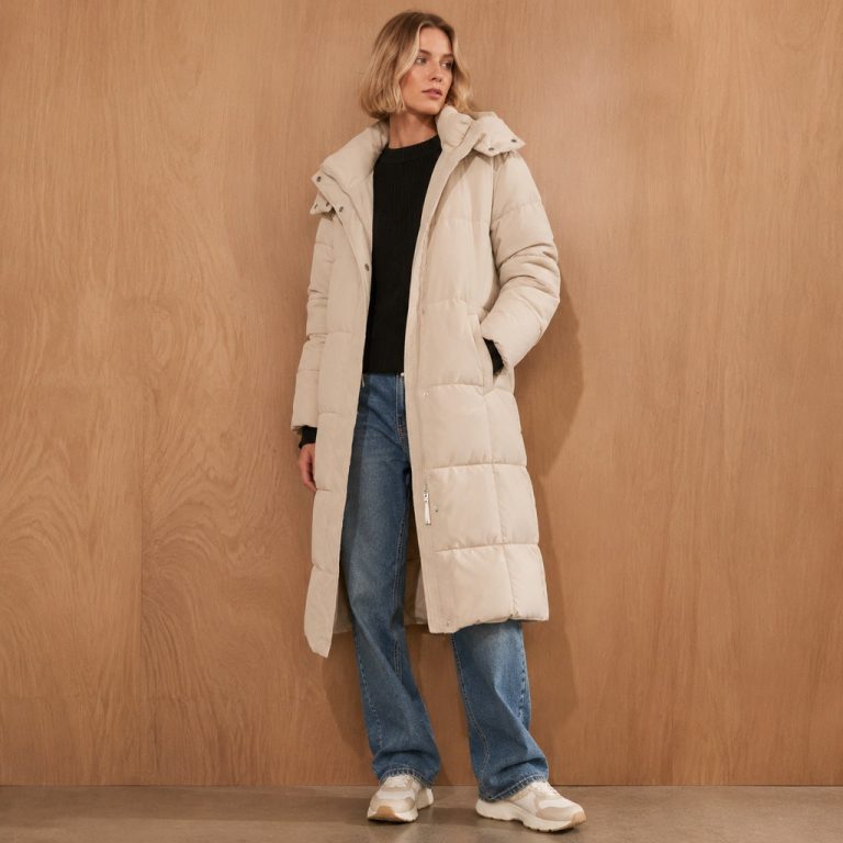 Women's cream longline puffer jacket model front view, full zip, drawcord hood with collar overlay, twin concealed hand pockets, side slits, mid-thigh length