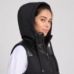 Women's black puffer vest model close-up hood shot, leather yoke trim, branded drawcord tips, two-way zip detail