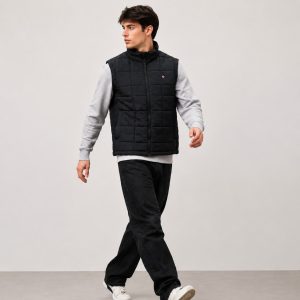 Men's black quilted vest model walking three-quarter view, full zip stand collar, side pockets, layered over grey sweatshirt, lifestyle studio