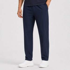 Male model wearing navy athletic workout pants with elastic waistband, right hand in side pocket, white sneakers, dark t-shirt, light gray studio backdrop