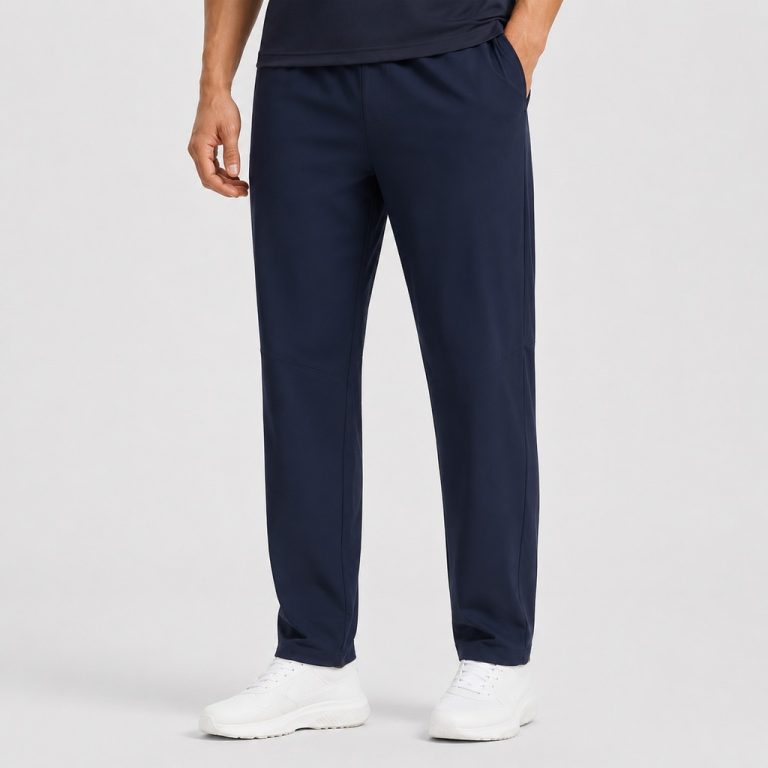 Male model wearing navy athletic workout pants with elastic waistband, right hand in side pocket, white sneakers, dark t-shirt, light gray studio backdrop