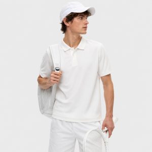 Male model wearing white short-sleeve polo workout shirt with 3-button placket, self-fabric collar, holding a tennis racket over right shoulder, white cap, white backpack, white shorts, light gray studio backdrop