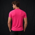 Back view of a man wearing a bright pink short-sleeve athletic t-shirt with small dots near the collar, paired with black shorts, photographed against a black studio backdrop.