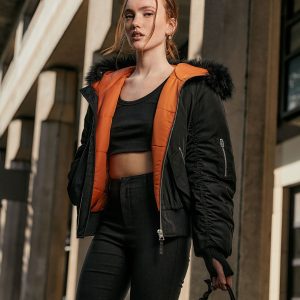 Female model wearing a black bomber jacket with orange quilted lining and faux fur-trimmed hood, paired with a black crop top and black jeans in an urban setting.