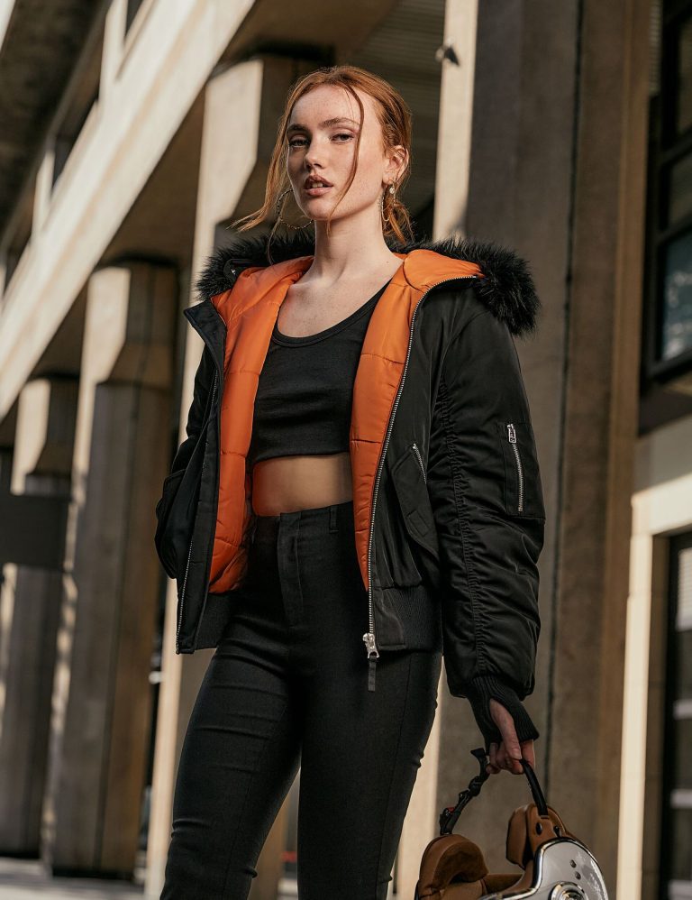 Female model wearing a black bomber jacket with orange quilted lining and faux fur-trimmed hood, paired with a black crop top and black jeans in an urban setting.