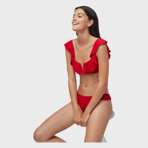 Model wearing a red bikini set with ruffle cap sleeves on the top, seated and smiling in a studio shot.