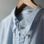 Close-up of a custom-made light blue shirt collar showing precise stitching and embroidered monogram detail on a wooden hanger