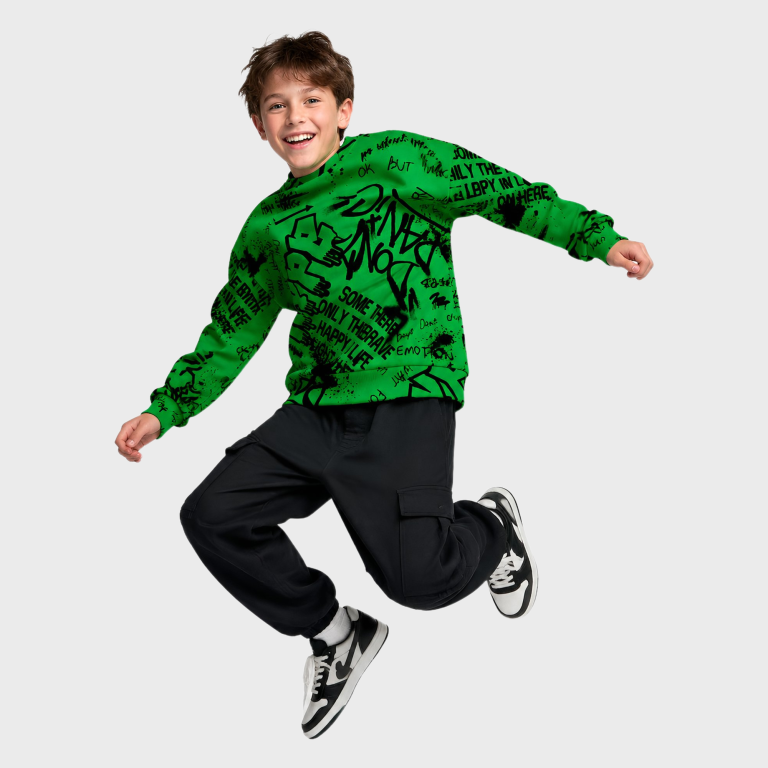 Smiling boy mid-jump wearing a green graffiti-print crewneck sweatshirt with black text, paired with black cargo joggers and sneakers, front view studio shot.