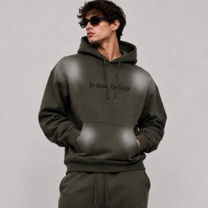 Men's olive green pullover hoodie with spray-print faded effect on shoulders and kangaroo pocket, "Be Good Do Right" gothic text printed in black on the chest center, drawcord hood, ribbed cuffs and hem, model front view wearing sunglasses, matching sweatpants