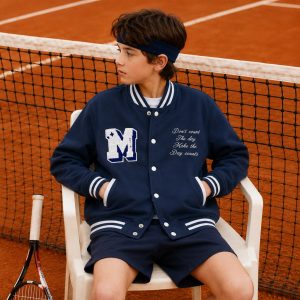 Boy's navy blue classic American varsity letterman bomber jacket with white chenille M letter patch on left chest and white cursive embroidered quote 'Don't count the day Make the Day counts' on right chest, white snap button front closure, ribbed collar cuffs and hem with white double stripe trim, two slanted side pockets with white trim, white contrast inner collar, kid model three-quarter view wearing navy headband white t-shirt under and navy shorts, clay tennis court background with editorial lifestyle composition