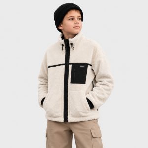 Boy's cream oatmeal sherpa fleece zip-up jacket with high funnel collar and black drawstring toggle, full-length black metal zipper front, black zippered chest pocket with small black patch with white text label inside (MA-1 style garment label 'RV CA CA 0000' + 'MADE IN CHINA', fully customizable per OEM buyer brand), two slanted welt side pockets at waist, contrasting black ribbed cuffs and hem, vertical black trim stripes along sleeve seams, kid model front three-quarter view wearing black knit beanie and khaki cargo pants, no external brand logos