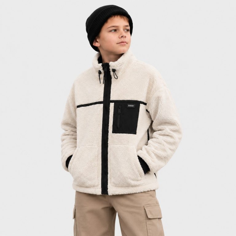Boy's cream oatmeal sherpa fleece zip-up jacket with high funnel collar and black drawstring toggle, full-length black metal zipper front, black zippered chest pocket with small black patch with white text label inside (MA-1 style garment label 'RV CA CA 0000' + 'MADE IN CHINA', fully customizable per OEM buyer brand), two slanted welt side pockets at waist, contrasting black ribbed cuffs and hem, vertical black trim stripes along sleeve seams, kid model front three-quarter view wearing black knit beanie and khaki cargo pants, no external brand logos