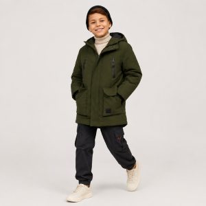 Boy's olive green mid-length utility parka puffer jacket with hood featuring black inner lining, full-length silver metal zipper front with hidden black snap button storm flap placket, vertical silver zip chest pocket on left chest, two large flap cargo pockets at waist with hidden snap button closures, small black rectangular MA-1 style garment label on lower right pocket (model code + MADE IN CHINA, fully customizable per OEM buyer), ribbed black knit cuffs, mid-length thigh-length silhouette, kid model front three-quarter view wearing black knit beanie cream turtleneck and black cargo pants with orange accents, clean light gray studio background
