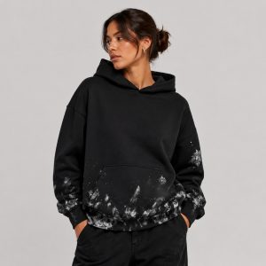 Women's oversized black pullover hoodie with abstract white-gray paint splatter and brushstroke distressing across the kangaroo pocket, hem, and sleeve cuffs, kangaroo pocket, drawcord hood, ribbed cuffs and hem, model front three-quarter view, low bun hairstyle, silver hoop earring