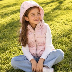Girl's light pink hooded puffer vest with full-length silver metal zipper front, scattered gold metallic five-point star print across the body, gathered elasticated waist, hood, paired with white long-sleeve knit top and light blue jeans, kid model sitting cross-legged on grass, smiling front view