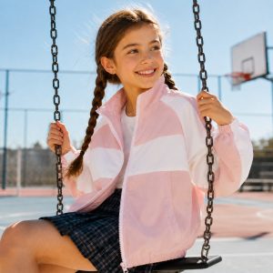 Girl's light pink and white color-block zip-up windbreaker with full-length silver metal zipper, stand collar, color-blocked panel construction with pink shoulders sleeves and lower body plus white chest stripe, elasticated cuffs and hem, kid model sitting on swing at outdoor playground, smiling front three-quarter view, braided hair, small stud earrings, plaid pleated skirt