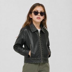 Girl's black studded leather (PU faux leather) jacket with full-length silver metal zipper front, point collar with silver studs along edges, two silver zip pockets at front waist, vertical silver stud lines along zipper placket, studded waistband and hem band, kid model front view wearing black heart-shape sunglasses, white tee under, hair down