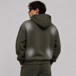 Men's olive green pullover hoodie with four symmetric faded spray-print spots on the upper-left, upper-right, lower-left, lower-right back area, drawcord hood, ribbed cuffs and hem, model back view wearing sunglasses, matching sweatpants