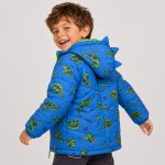 Boy's royal blue diamond quilted puffer jacket back view, hood up with 3D fabric dinosaur spike/horn on hood edge, allover scattered cartoon dinosaur silhouette print and 'WILD ONE' slogan and lightning bolt decorations continuing across back, lime green drawstring toggle at hem, contrasting lime green inner lining visible at hood, ribbed cuffs and hem, kid model back view brown hair, black pants