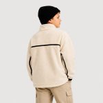 Boy's cream oatmeal sherpa fleece zip-up jacket back view, high funnel collar, horizontal black trim stripe across upper back at chest level, vertical black trim stripes along sleeve seams, contrasting black ribbed cuffs and hem, kid model back view with black knit beanie and khaki cargo pants, no external brand logos or text