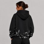 Women's oversized black pullover hoodie with abstract white-gray paint splatter and brushstroke distressing continuing across the back hem, lower sleeves, and side body, kangaroo pocket, drawcord hood down, ribbed cuffs and hem, model back view, low bun hairstyle, silver hoop earring