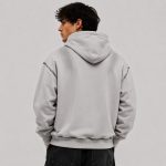 Men's oversized light gray pullover hoodie with kangaroo pocket, drawcord hood, exposed raw-edge distressed seams continuing on back shoulder drop-line and bottom hem ribbing, ribbed cuffs and hem, model back view, silver hoop earring