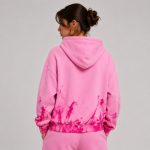 Women's light pink zip-up hoodie with drawcord hood, abstract magenta and hot pink paint splatter effect continuing across back hem and lower sleeves, ribbed cuffs and waistband, model back view with low bun hairstyle, silver hoop earring