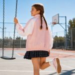 Girl's light pink and white color-block zip-up windbreaker back view, full back panel pink with white sleeves, stand collar, elasticated hem, no text or logo, kid model on swing at outdoor playground, braided hair, plaid pleated skirt