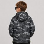 Boy's dark gray black and white abstract geometric halftone camo print hooded windbreaker flat lay product photo front, hood up with neon yellow green drawstring, full-length silver zipper, two slanted side pockets, ribbed elasticated cuffs, toggle hem drawcord with neon yellow cord ends, clean light gray background for B2B catalog, no text or logo