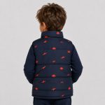 Boy's navy blue puffer vest flat lay product photo front, high funnel collar with contrasting orange inner lining visible, full-length silver metal zipper front, two slanted side pockets, allover scattered orange dinosaur silhouette print, clean light gray background for B2B catalog, no text or logo
