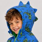 Boy's royal blue diamond quilted puffer jacket closeup view with hood pulled up over face, lime green metal zipper pulled up, contrasting lime green inner lining visible at hood interior, allover scattered cartoon dinosaur print and 'WILD ONE' slogan and lightning bolt decorations visible, kid model looking at camera smiling with eyes visible
