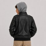 Boy's black faux leather (PU leather) bomber jacket flat lay product photo front, heather gray hooded sweatshirt worn underneath with hood up visible at neckline, full-length silver metal zipper, two chest flap pockets with silver snap button closures, two slanted welt side pockets, epaulettes on each shoulder with silver snap button, ribbed cuffs and hem, no text or logo, clean white studio background for B2B catalog