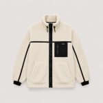 Boy's cream oatmeal sherpa fleece zip-up jacket flat lay product photo front, high funnel collar with black drawstring toggle, full-length black metal zipper, black zippered chest pocket with small black patch with white text MA-1 style garment label inside (fully customizable), two slanted welt side pockets, contrasting black ribbed cuffs and hem, vertical black trim stripes along sleeve seams, clean white studio background for B2B catalog