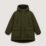 Boy's olive green mid-length utility parka puffer jacket flat lay product photo front, hood with drawstring and black inner lining visible, full-length silver metal zipper front with hidden black snap button storm flap placket, vertical silver zip chest pocket on left chest, two large flap cargo pockets at waist with hidden snap button closures, small black rectangular MA-1 style garment label on lower right pocket, ribbed black knit cuffs visible at sleeve ends, mid-length thigh-length silhouette, clean light gray studio background for B2B catalog