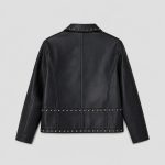 Girl's black studded leather (PU faux leather) jacket flat lay product photo front, full-length silver metal zipper, point collar with studs along collar points, vertical silver stud lines along entire front zipper placket, two silver zip pockets at front waist, studded waistband and hem band, clean light gray background for B2B catalog