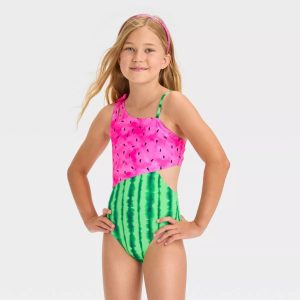 Young girl modeling a children's one-piece swimsuit with a pink watermelon-patterned top and green striped watermelon bottom, posing with hands on hips in a studio setting.