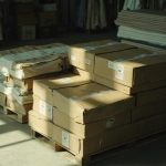 Neatly stacked cartons of finished garments on pallets inside a clean apparel factory warehouse, with fabric rolls on racks in the background under soft natural light.