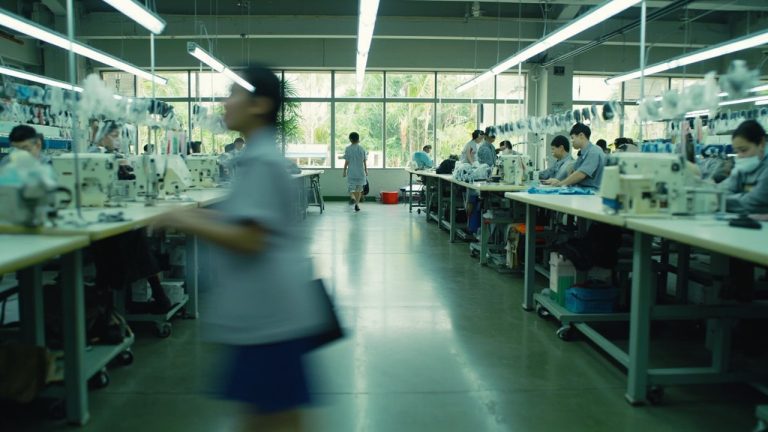 Industrial apparel production line with multiple workers operating sewing machines under fluorescent lights in a spacious facility.